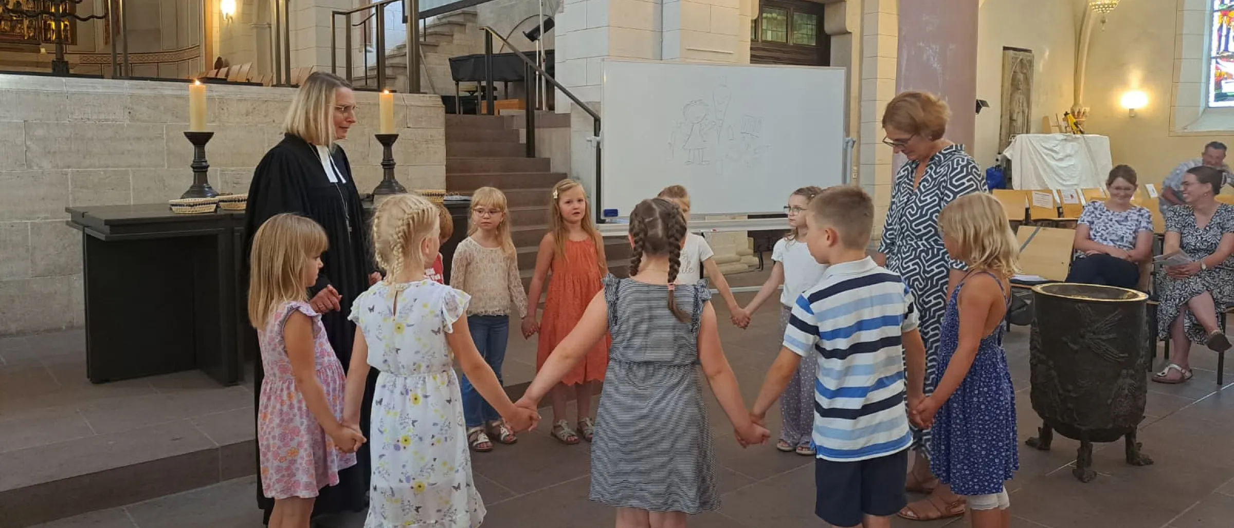 Zu sehen sind die Schülerinnen und Schüler der 1a, die einen Segenskreis bilden und sich an den Händen halten. Sie stehen vor dem Altar, mit dabei sind die Klassenlehrerin Christiane Lillig-Konerding und Pröpstin Meike Bräuert-Ehgart